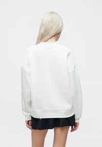 Balta pārstrādāta krekla džemperis ar apaļu kaklu, nolaižamām plecu daļām un ribotām piedurknēm. Gluda tekstūra, minimālisma dizains, valkāts virs melnas krokotās svārku.