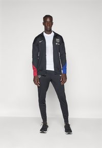 Nike sportjakke i sort med røde og blå accenter, parret med matchende sorte bukser og sneakers, med et logo på brystet.