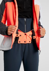 Sportjacke in Orange und Schwarz, mit einem Reißverschluss vorne und einer darunter sichtbaren, enganliegenden schwarzen Basisschicht. Druckknöpfe im Taillenbereich.