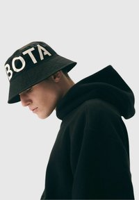 Czarny kapelusz typu bucket z dużymi, białymi literami "BOTA", zestawiony z czarną bluzą z kapturem. Kapelusz ma szeroki rondo i teksturowany materiał.