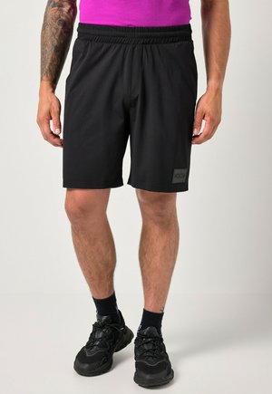 Mand iført sorte atletikshorts, sorte sokker, sorte sneakers og en lilla skjorte, står mod en lys baggrund.