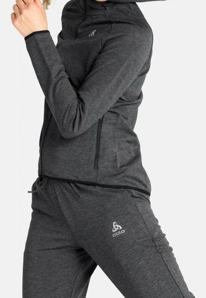 Persoon met donkergrijze hoodie met rits en bijpassende joggingbroek met Odlo-logo, staand met één arm gebogen bij het gezicht.