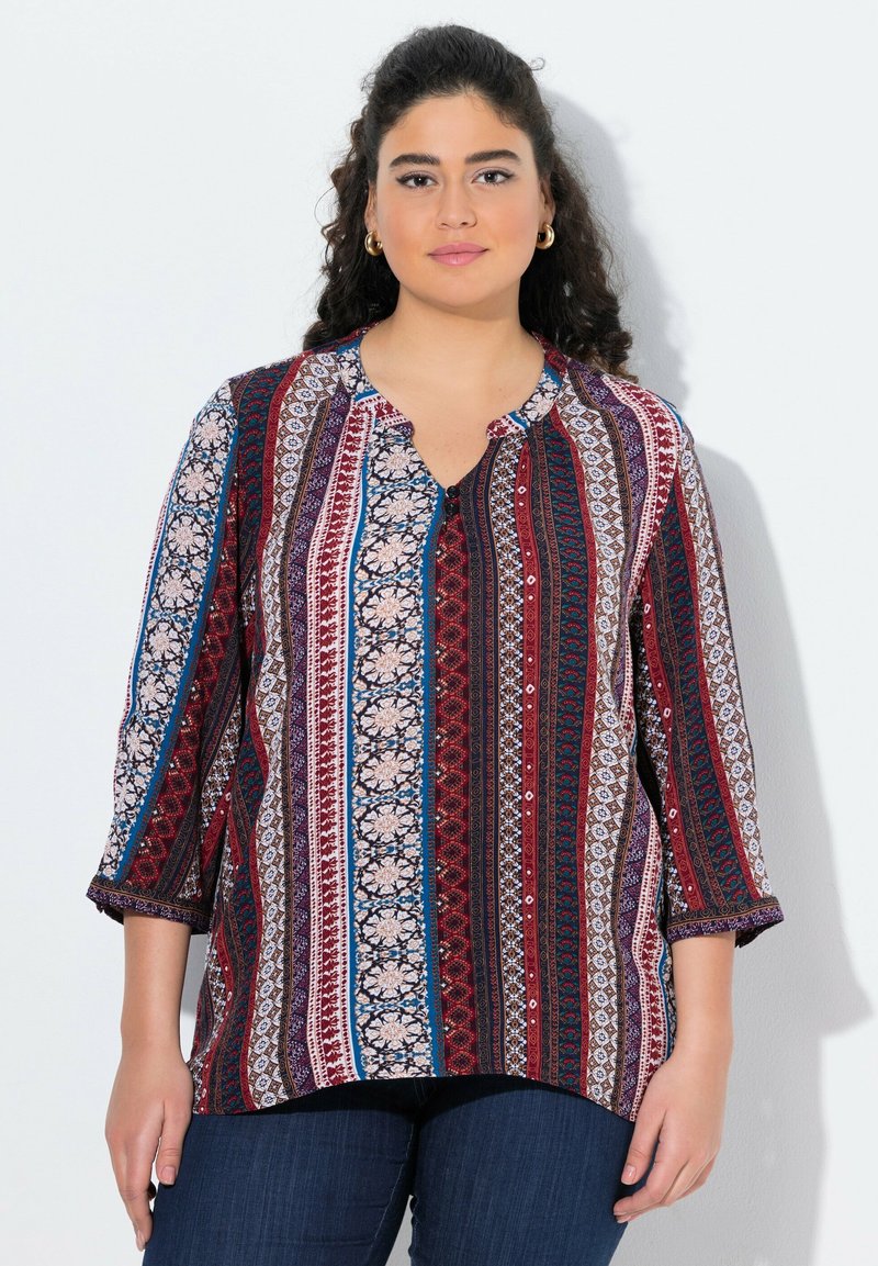Blusa multicolore con motivi floreali e geometrici, scollatura rotonda, dettagli con bottoni e maniche a tre quarti. Il tessuto ha una texture liscia.