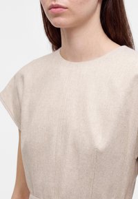 Gros plan d'une personne portant une robe beige à manches courtes texturée avec un col rond et de longs cheveux bruns.