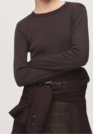 Personne portant un haut manches longues marron foncé avec les bras croisés, une veste assortie nouée à la taille, et un pantalon à carreaux visible en dessous.