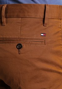 Pantaloni in cotone marroni con una tasca posteriore con un bottone nero e un piccolo logo ricamato in rosso, bianco e blu vicino alla tasca.