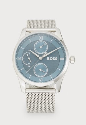Montre en maille argentée avec un cadran rond bleu, des chiffres romains, trois sous-cadrans et une surface texturée. Le nom de la marque "BOSS" est affiché de manière proéminente.