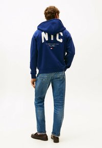 Felpa blu navy con grande scritta bianca "NYC" sul retro, dotata di cappuccio con lacci e polsini a coste, abbinata a jeans azzurri.