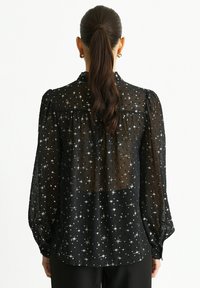 Black sheer blouse with long puff sleeves, adorned with silver star patterns, showing a gathered back yoke and buttoned cuffs.
