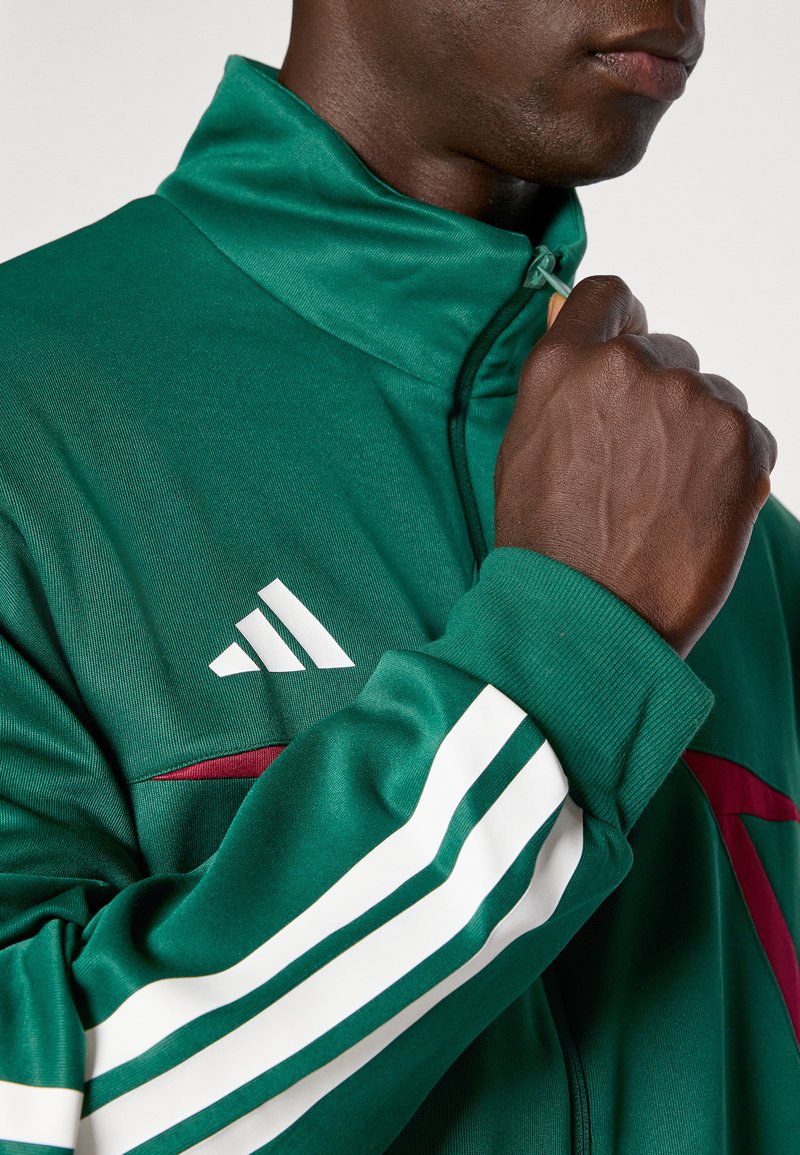 Groene sportjacket met een opstaande kraag, witte strepen op de mouwen, bordeauxrode accenten en een wit Adidas logo op de borst.