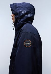 Námořnická modrá bunda s kapucí, vybavená nášivkou s logem na rukávu, stahovacími šňůrami a hladkou texturou. Zobrazena boční část.