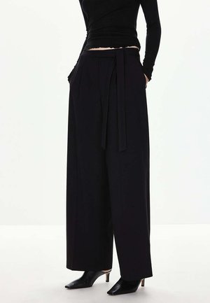 Femme portant un pantalon noir taille haute à jambes larges avec une ceinture en tissu, associé à des chaussures noires à talons pointus et un haut noir moulant.