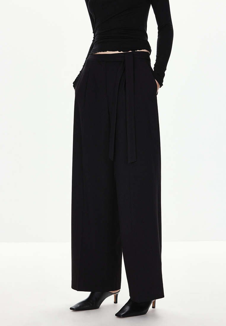 Femme portant un pantalon noir taille haute à jambes larges avec une ceinture en tissu, associé à des chaussures noires à talons pointus et un haut noir moulant.