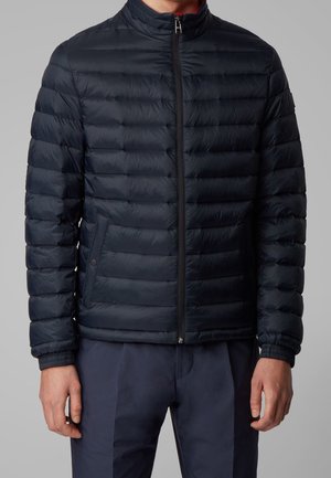 Man wearing a navy blue quilted zip-up jacket with side pockets and navy trousers, standing against a gray background.