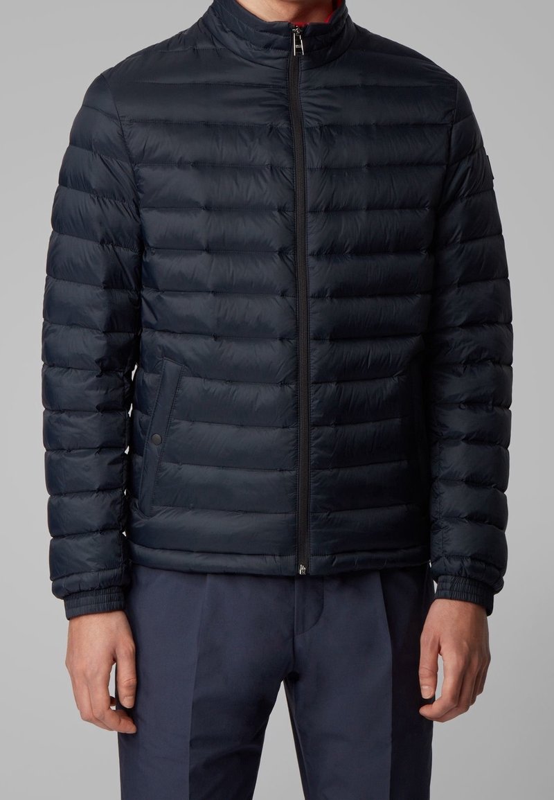 Homme portant une veste matelassée bleue marine zippée avec poches latérales et un pantalon bleu marine, debout devant un fond gris.