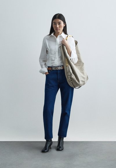 Camisa blanca de botones con pliegues, pantalones vaqueros de mezclilla azules, botas negras hasta el tobillo y un bolso de hombro beige claro con acentos dorados y marca.
