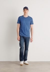 Μπλε κοντομάνικη μπλούζα, σκούρα τζιν και λευκά αθλητικά. Το μοντέλο φοράει ένα σκούρο καπέλο μπέιζμπολ. Απλός σχεδιασμός, μονόχρωμα, χωρίς μοτίβα.