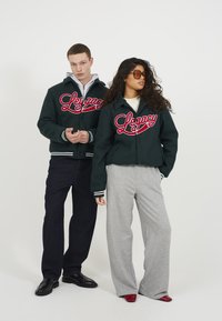 Mørkegrønne zip-up jakker med "Legacy" i røde bogstaver, grå sweatpants og kontrastfuldt fodtøj; modellen poserer mod en ensfarvet baggrund.