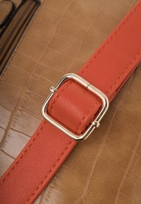 Bracelet en cuir orange avec boucle carrée dorée sur surface en cuir brun texturé, montrant des coutures détaillées et des motifs de grain.