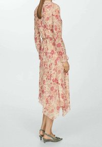 Robe fleurie en chiffon doux, présentant une base pastel avec des fleurs roses, des manches longues, une taille cintrée et un ourlet asymétrique. Associ ée à des talons verts.