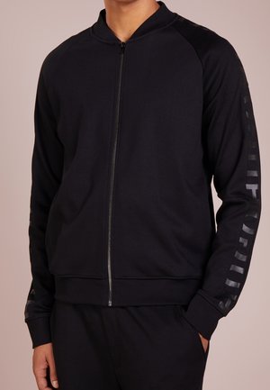 Giacca nera con zip realizzata in tessuto morbido, caratterizzata da maniche raglan, polsini a coste e una stampa logo testurizzata lungo il braccio sinistro.