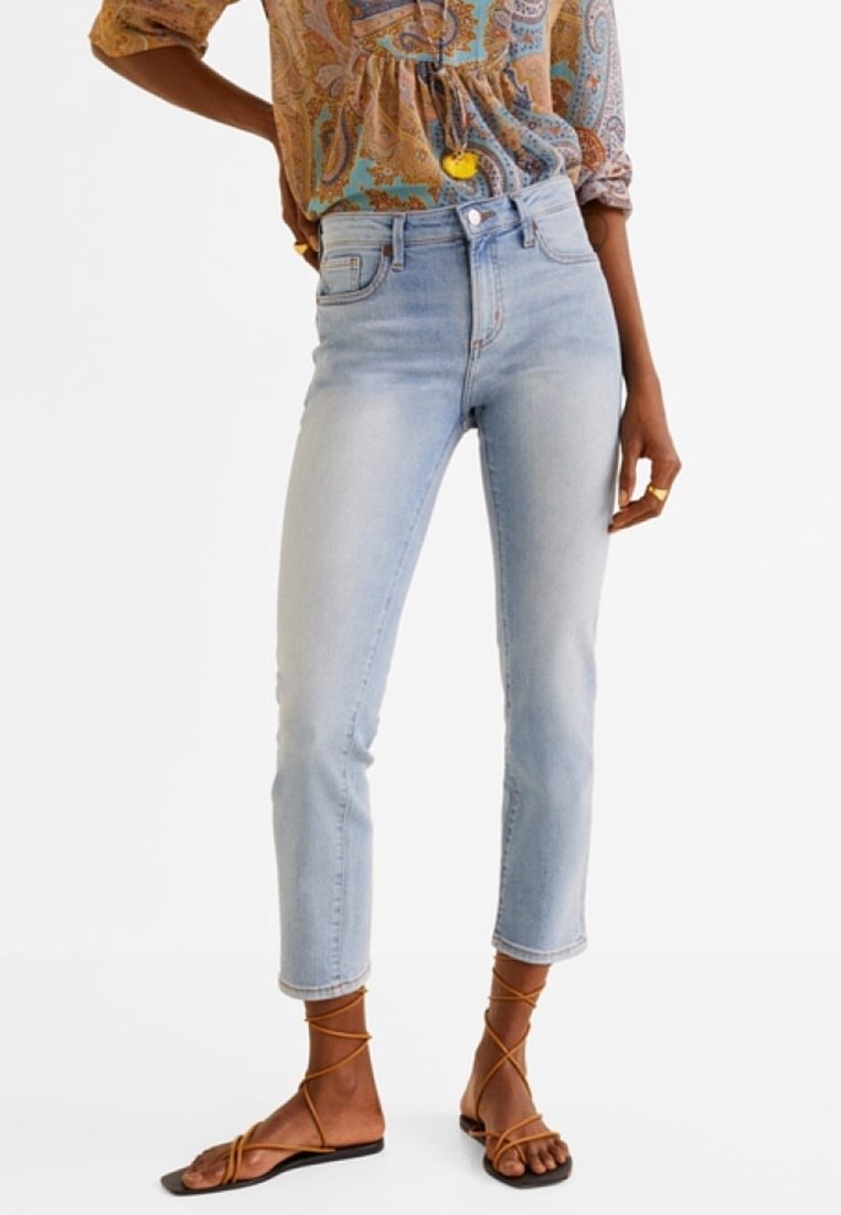 Personne portant un jean court bleu clair, un blouse à manches longues avec un motif, et des sandales plates brunes à lanières, se tenant avec la main sur la hanche.