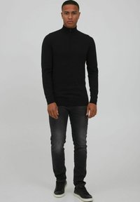 Schwarzes Langarmshirt mit Reißverschluss und aus weichem Stoff, kombiniert mit dunkelgrauen Jeans und schwarzen Schuhen mit weißen Sohlen.