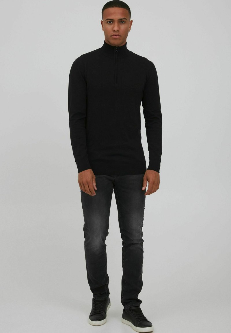 Schwarzes Langarmshirt mit Reißverschluss und aus weichem Stoff, kombiniert mit dunkelgrauen Jeans und schwarzen Schuhen mit weißen Sohlen.