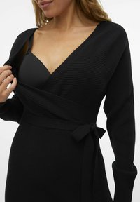 Abito avvolgente nero in tessuto a coste, con scollatura a V profonda, maniche lunghe e una cintura in vita per la regolazione. Silhouette liscia e aderente.