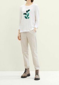 Maglietta bianca a maniche lunghe con grafica di una foglia verde, abbinata a pantaloni leggeri beige e stivali marroni con lacci e suola spessa.