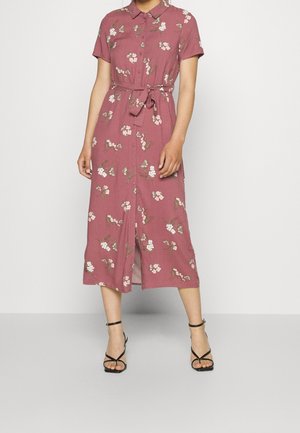 Frau in einem mauvefarbenen, blumigen Midikleid mit kurzen Ärmeln, Knopfleiste, gebundenem Taillenbund und schwarzen, schmalen, hochhackigen Sandalen.