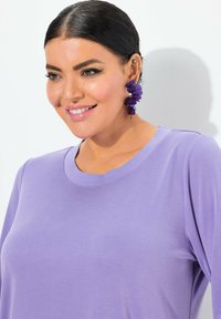 Lila Kurzarmshirt mit Rundhalsausschnitt, das eine glatte Textur aufweist. Auffällige lila Statement-Ohrringe sorgen für visuelles Interesse.