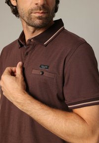 Polo marron avec col texturé, détail de poche sur bouton et accents de rayures contrastants. Le modèle se tient les mains sur la poitrine, mettant en valeur le design.