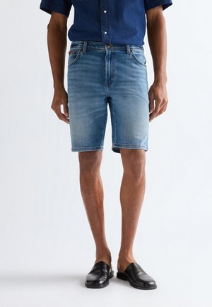 Mężczyzna w wyblakłych, niebieskich szortach dżinsowych, ciemnoniebieskiej koszuli z krótkim rękawem i czarnych butach wsuwanych, stojący na tle białego tła.