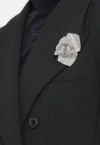 Blazer nero con una spilla a forma di fiore in strass argentati sulla giacca. Tessuto strutturato e dettaglio con un singolo bottone nero.