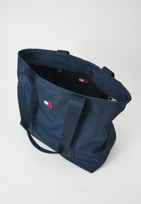 Geantă de umăr de culoare bleumarin, cu două mânere rezistente, un buzunar cu fermoar în interior și un patch cu logo din material textil. Material din nailon texturat cu formă structurată.