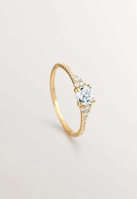 Anillo de oro con una gema central de color azul claro, flanqueada por dos piedras claras más pequeñas a cada lado, y una banda engastada con diamantes.