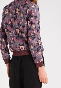 Kurtka bombka w kwiaty z fioletowego materiału, z różowymi i czarnymi wzorami kwiatowymi, ściągaczami na rękawach oraz teksturowanym wykończeniem.