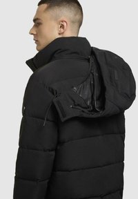 Schwarze Puffjacke mit hohem Kragen, Reißverschluss und Klettverschluss-Details sowie einer strukturierten Oberfläche. Mit abnehmbarem Kapuzen und sichtbarem Futter.