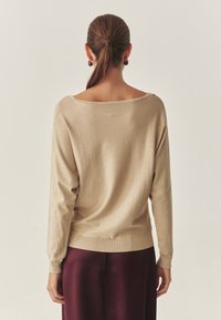 Bež dolg rokav pulover z razširjenim izrezom in rebrastimi manšetami. Mehka tekstura, nošena čez temno bordo oblačilo. Prijeten kroj, minimalističen dizajn.