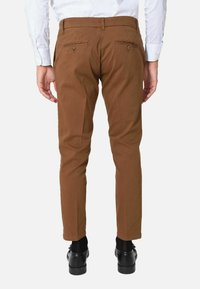 Pantaloni in cotone marroni con vestibilità slim, dotati di due tasche posteriori con bottone e una texture liscia, indossati con scarpe nere.