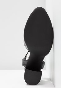 Semelle de chaussure noire avec talon texturé et bout pointu, équipée d'une bride autour de la cheville, reposant contre une surface blanche.