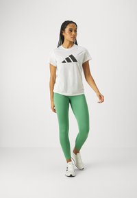 Hvid T-shirt med sort Adidas-logo, sammen med tætsiddende grønne leggings og hvide atletiske sko, mod en simpel grå baggrund.