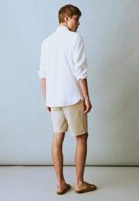 Chemise blanche à manches longues, short beige, sandales marron. Design simple, couleurs claires, texture lisse, détail de poignet visible et vue de dos.