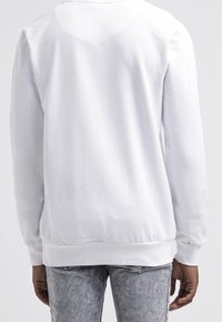 Bílá mikina s kulatým výstřihem, dlouhými rukávy a žebrovanými lemy na rukávech a spodním okraji. Měkká látka s hladkou texturou, bez viditelných vzorů.