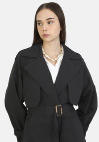 Black trench coat with a wide collar, belted waist, and shoulder accents, worn over a white shirt, complemented by a large pearl necklace.