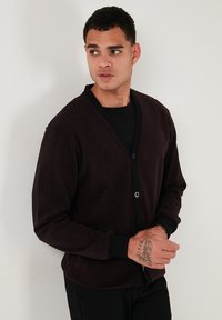 Cardigan marrone scuro con scollo a V, chiusura con bottoni e contrasti neri alle maniche e all'orlo. Tessuto morbido e di peso medio.