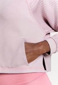 Main foncée d'une personne dans la poche latérale d'une veste rose clair avec des manches matelassées, au-dessus d'un pantalon rose assorti.