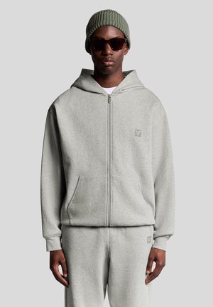 Un hoodie zippé gris en tissu doux, avec une poche kangourou, des poignets côtelés et un logo brodé sur la poitrine. Associé à un pantalon assorti.