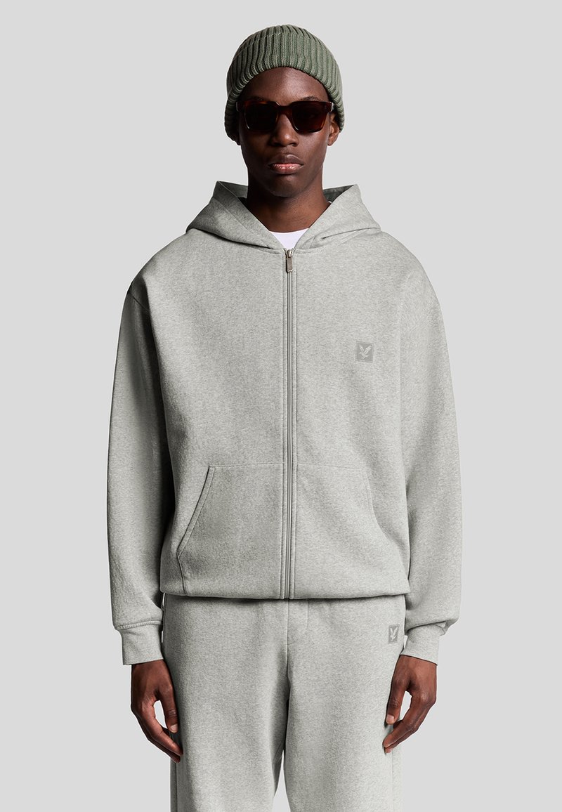 Grå zip-up hoodie lavet af blød stof har en kengurulomme, ribbede ærmer og et broderet logo på brystet. Matchet med matchende bukser.